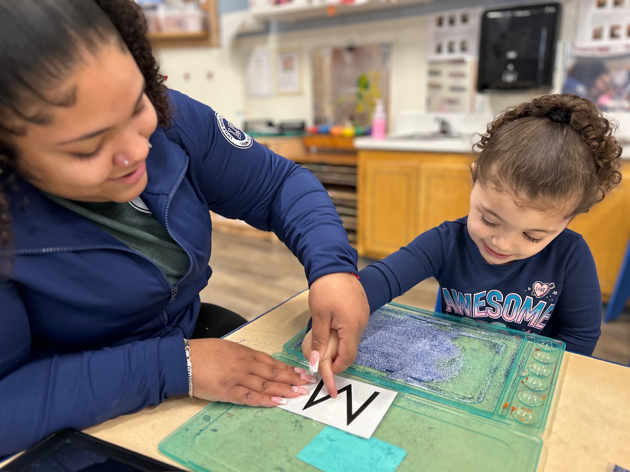 An LPC teacher guiding a young girl as she traces letters using a sensory tray in the classroom